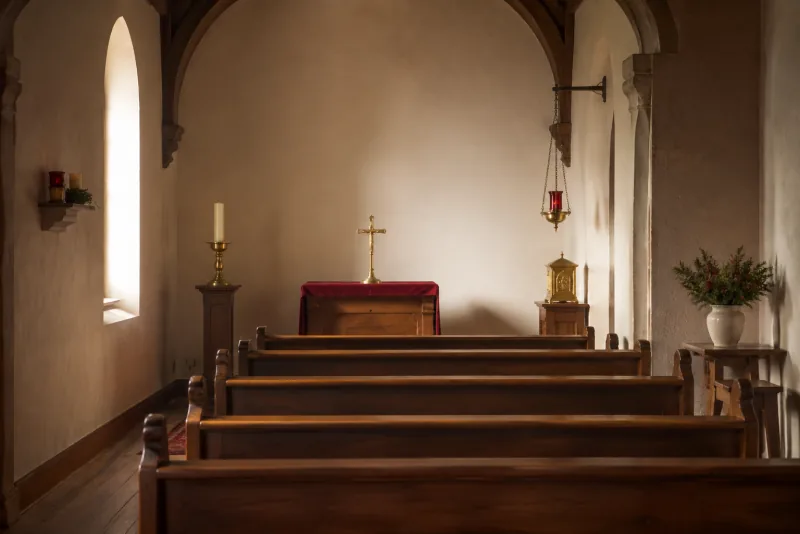 Rows of wooden pews face a small gold crucifix and a red-covered altar in a quiet chapel.