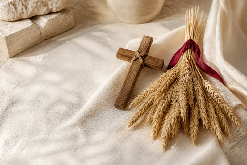 A twine-bound wooden cross lies beside a sheaf of ripe wheat tied with red ribbon on ivory linen.