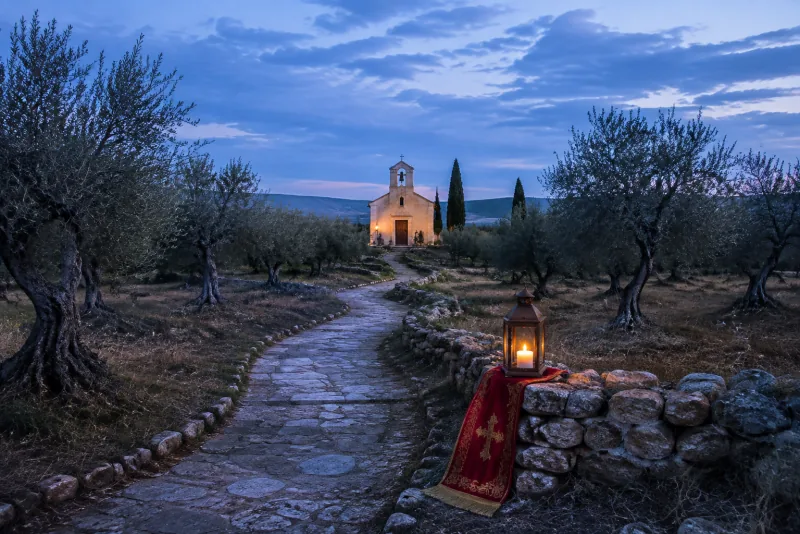 A stone path winds through an olive grove toward a small chapel at blue hour, with a glowing lantern on a red cloth.