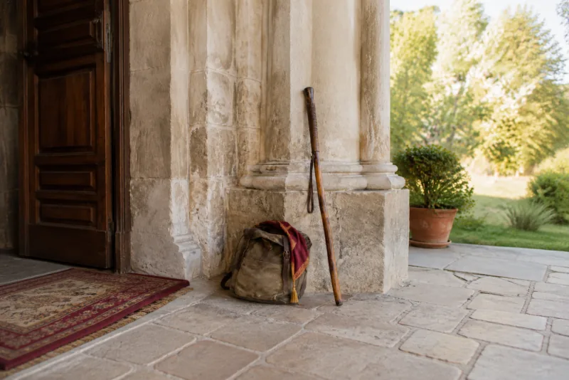 A wooden walking staff and canvas satchel rest beside a stone church doorway.