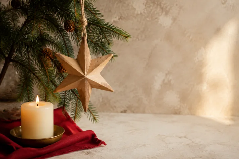 A wooden star hangs among evergreen branches beside a glowing candle.